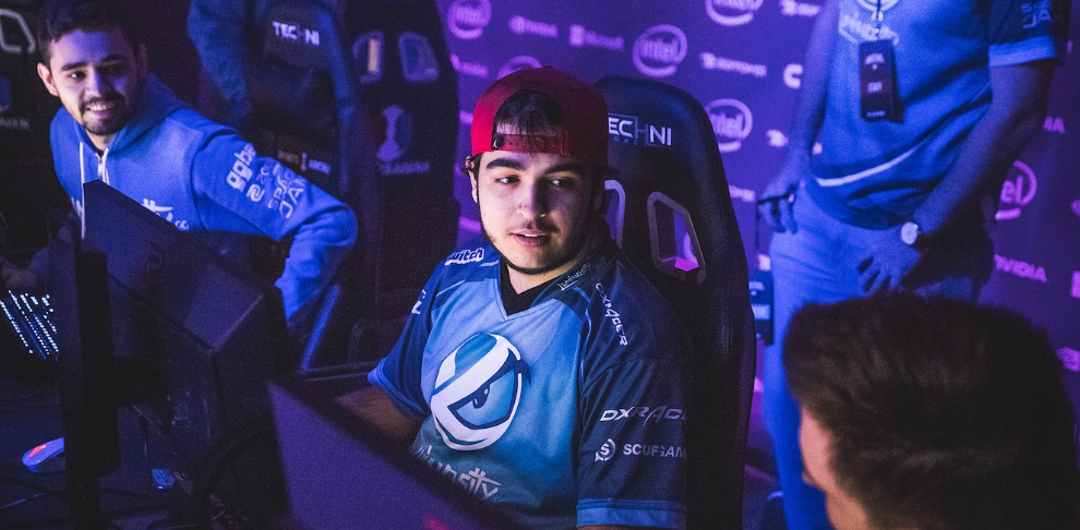 Chelo de bone para trás em cadeira gamer em frente a setup com companheiros de time