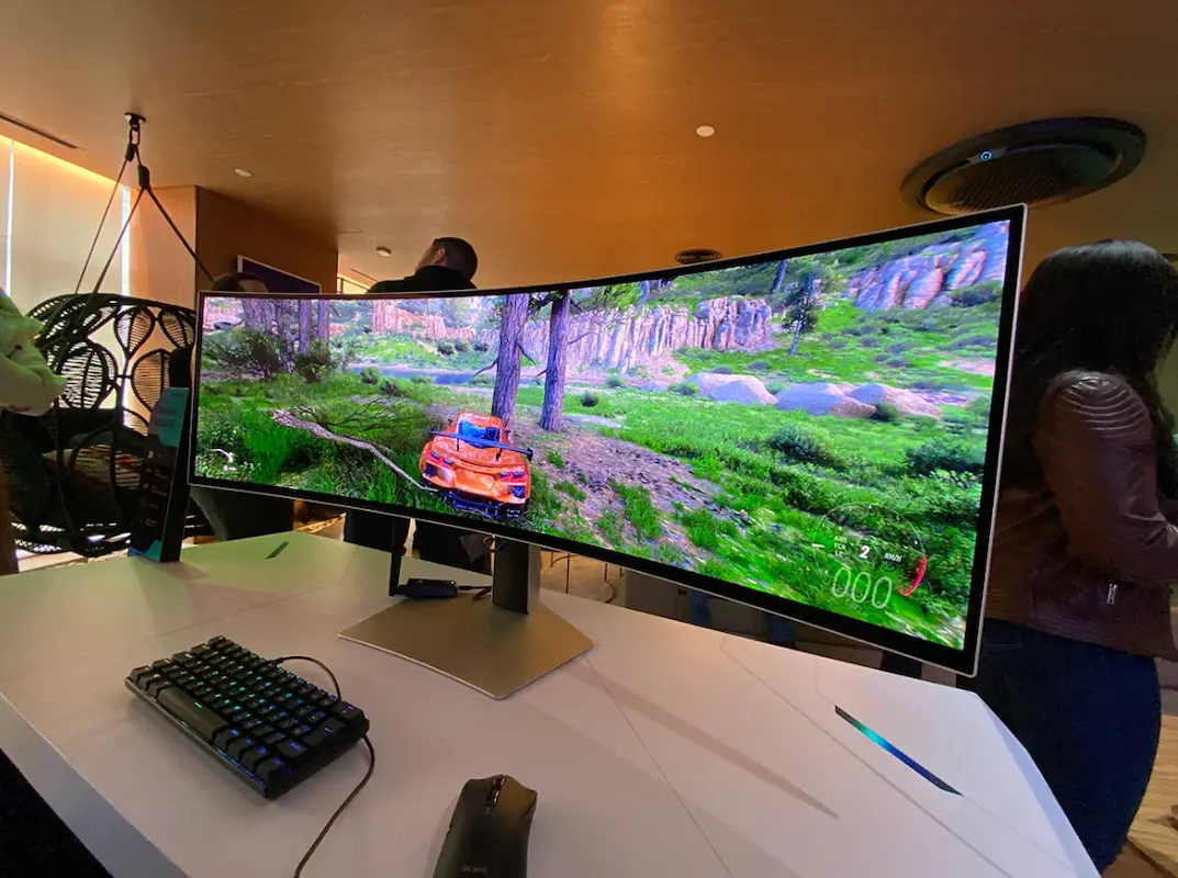 Demonstração de monitor curvo ultra-wide em ângulo lateral, exibindo jogo de corrida com gráficos realistas, teclado mecânico e mouse gamer em uma mesa branca.
