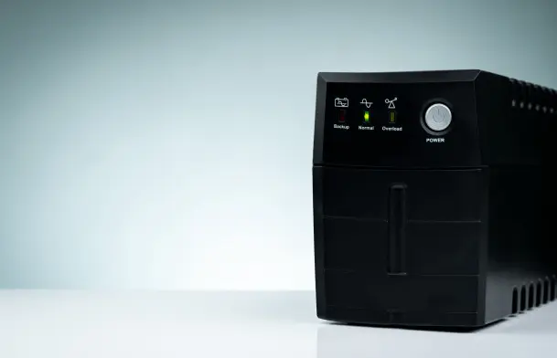 Nobreak preto sobre uma superfície branca, com luzes indicadoras acesas no painel frontal mostrando os modos Backup, Normal e Overload, além de um botão de energia prateado.